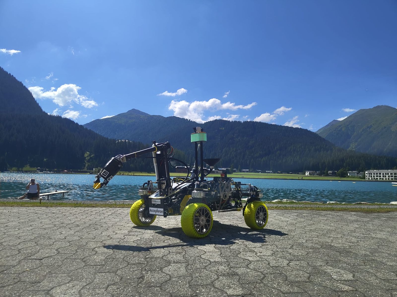 A Mars rover with yellow wheels and a raised robotic arm, parked on a promenade beside Lake Davos, forested mountains behind it.